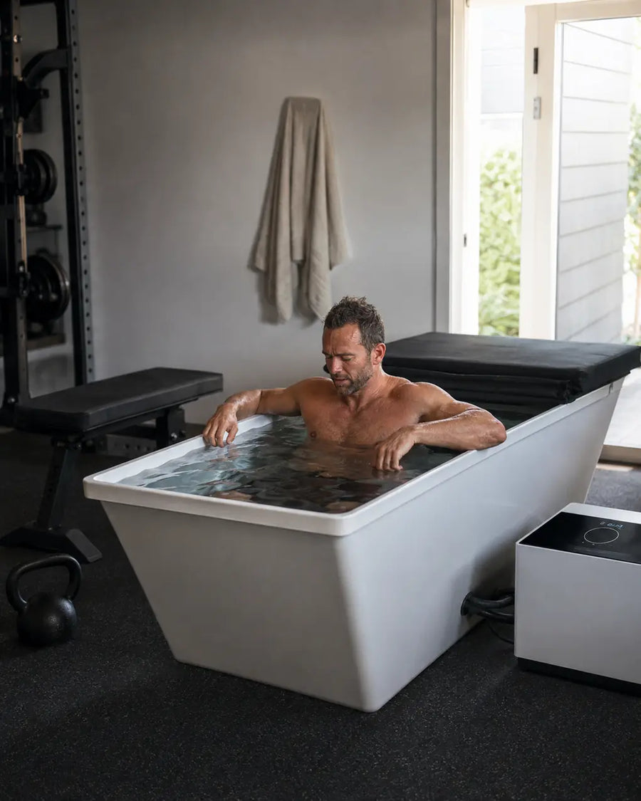 Man seated in a full length cold plunge tub beside workout equipment in a home gym.